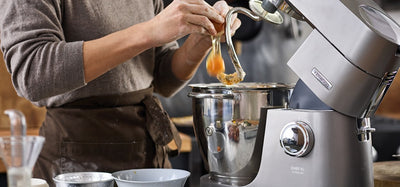 A person using a Kenwood stand mixer with ingredients on a kitchen counter.